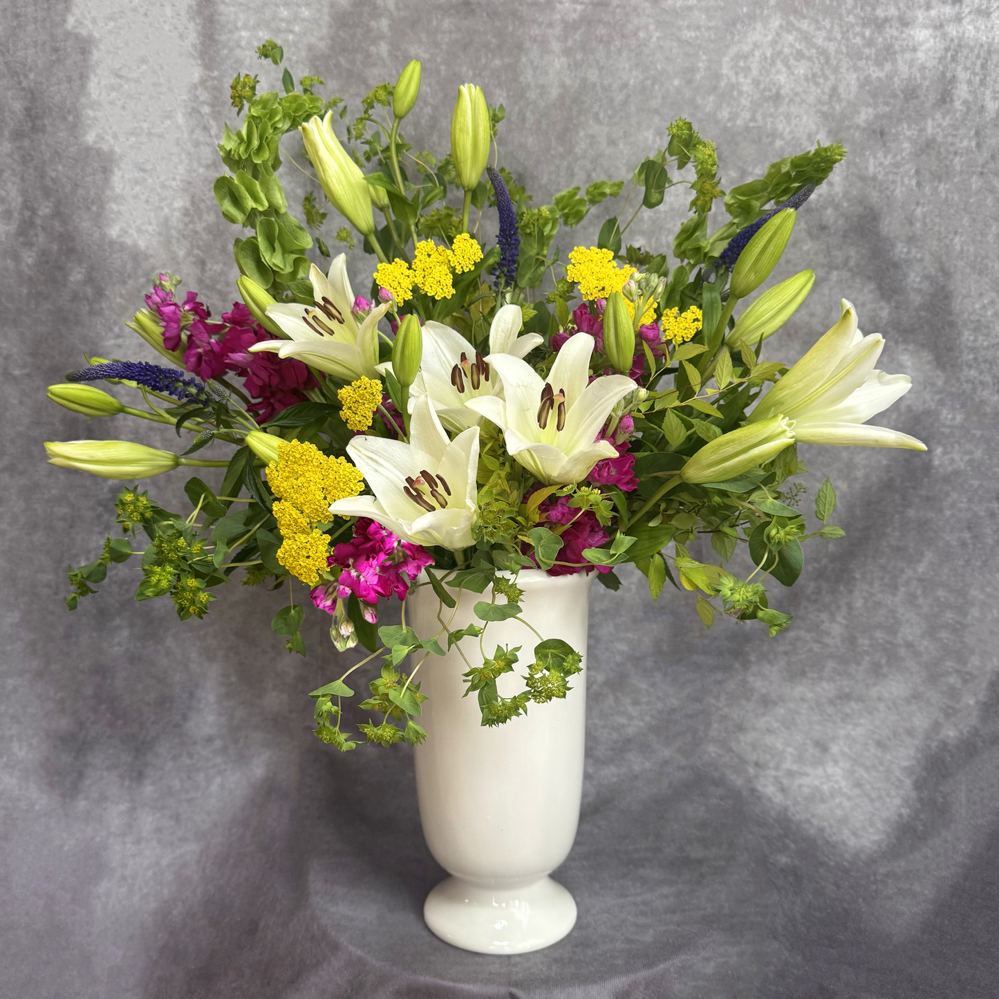 Floral arrangement with white lilies, yellow flowers, and greenery in a white vase on a gray background